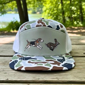 May include: A camouflage-patterned baseball cap with a white mesh back and a flat brim. The front panel features an embroidered image of a dog chasing a rabbit. The cap's design incorporates shades of green, brown, and blue.