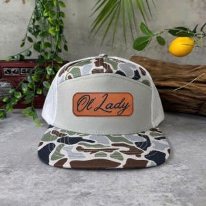 May include: A camouflage print trucker hat with a white mesh back and a light gray front panel. The front panel features a brown leather patch with the words "Ol Lady" in a cursive font.