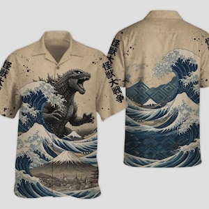 May include: A beige short-sleeve button-down shirt featuring a graphic of a dark grey monster emerging from blue ocean waves, a snow-capped mountain, and Japanese text. The design includes black ink splatters and a repeating pattern of waves.