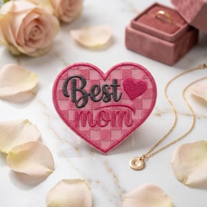 May include: A pink heart-shaped embroidered patch with the words "Best Mom" in black script and a small pink heart. The patch has a checkered pattern and is surrounded by rose petals, a gold necklace, and a ring box.