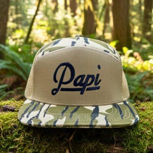 May include: A khaki-colored trucker hat with a mesh back and a camouflage brim. The front panel features the word "Papi" in navy blue script. The hat is set against a backdrop of a forest.