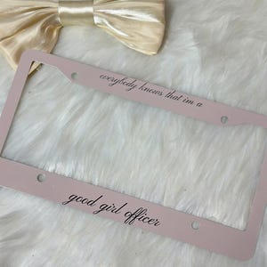 May include: A light pink license plate frame with the text "everybody knows that I'm a good girl officer" in black script. A cream-colored bow is in the background.