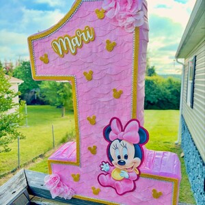 May include: A number one shaped pi&ntilde;ata in light pink with a scalloped texture, gold glitter trim, and small gold glitter mouse head silhouettes. It features the name Mari in gold script, a pink flower cluster, and a cartoon mouse character.