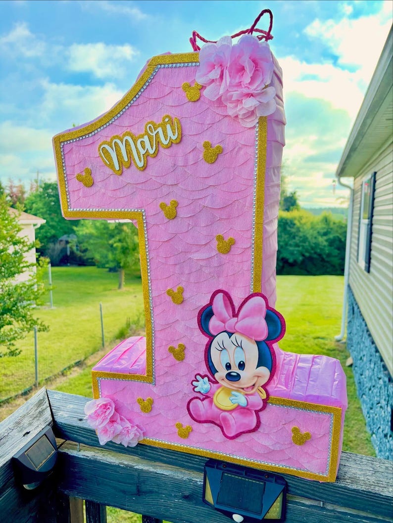 May include: A number one shaped pi&ntilde;ata in light pink with a scalloped texture, gold glitter trim, and small gold glitter mouse head silhouettes. It features the name Mari in gold script, a pink flower cluster, and a cartoon mouse character.