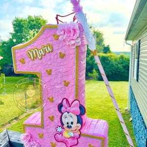 May include: A pink number one shaped pi&ntilde;ata featuring a scalloped texture, gold glitter trim, and small gold mouse head silhouettes. It displays the name Mari in gold script, a Minnie Mouse graphic, pink fabric flowers, and a matching pink stick.