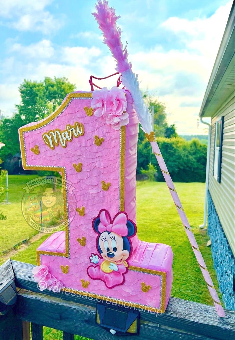 May include: A pink number one shaped pi&ntilde;ata featuring a scalloped texture, gold glitter trim, and small gold mouse head silhouettes. It displays the name Mari in gold script, a Minnie Mouse graphic, pink fabric flowers, and a matching pink stick.