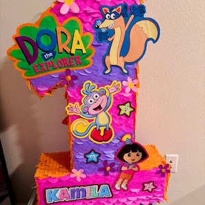 Peut inclure: Une piñata en forme de chiffre un décorée de franges en papier rose et violet. Elle présente des découpes de Dora l'exploratrice, Babouche le singe et Chipeur le renard. Le texte Dora the Explorer et le prénom Kamila sont imprimés devant.
