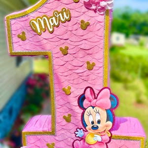 May include: A pink number one shaped pinata featuring a scalloped paper texture, gold glitter trim, and small gold glitter mouse head silhouettes. The name Mari is written in white script, and a cartoon mouse character is attached to the front.