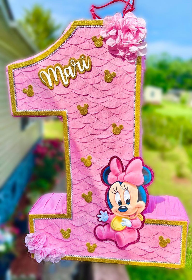 May include: A pink number one shaped pinata featuring a scalloped paper texture, gold glitter trim, and small gold glitter mouse head silhouettes. The name Mari is written in white script, and a cartoon mouse character is attached to the front.