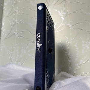 May include: A navy blue book with the title "coraline" in silver lettering on the spine. The cover features a spiderweb design. Two white buttons are visible on the spine. The book is resting on a white, textured surface.