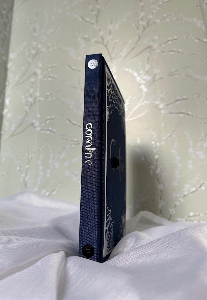 May include: A navy blue book with the title "coraline" in silver lettering on the spine. The cover features a spiderweb design. Two white buttons are visible on the spine. The book is resting on a white, textured surface.