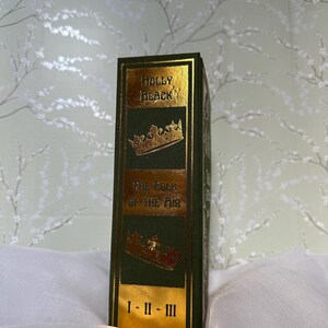 May include: A hardcover book with a dark green cover and gold lettering. The book's spine features the title "HOLLY BLACK" and "THE FOLK OF THE AIR" with gold crown illustrations. The book's volume numbers are indicated at the bottom.