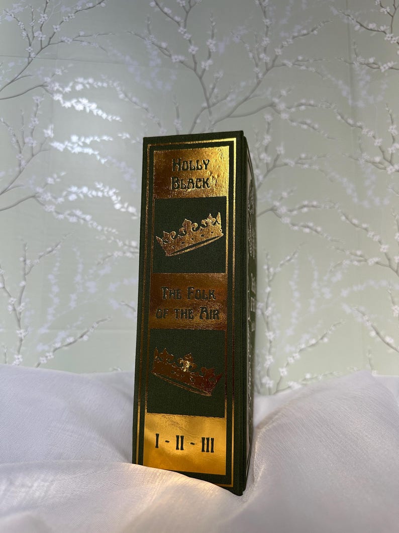 May include: A hardcover book with a dark green cover and gold lettering. The book's spine features the title "HOLLY BLACK" and "THE FOLK OF THE AIR" with gold crown illustrations. The book's volume numbers are indicated at the bottom.