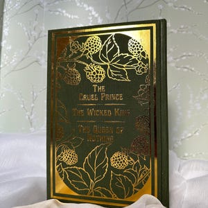 May include: A hardcover book with a dark green cover and gold foil accents. The title "The Cruel Prince," "The Wicked King," and "The Queen of Nothing" are displayed in gold lettering. The cover features gold illustrations of berries and leaves.