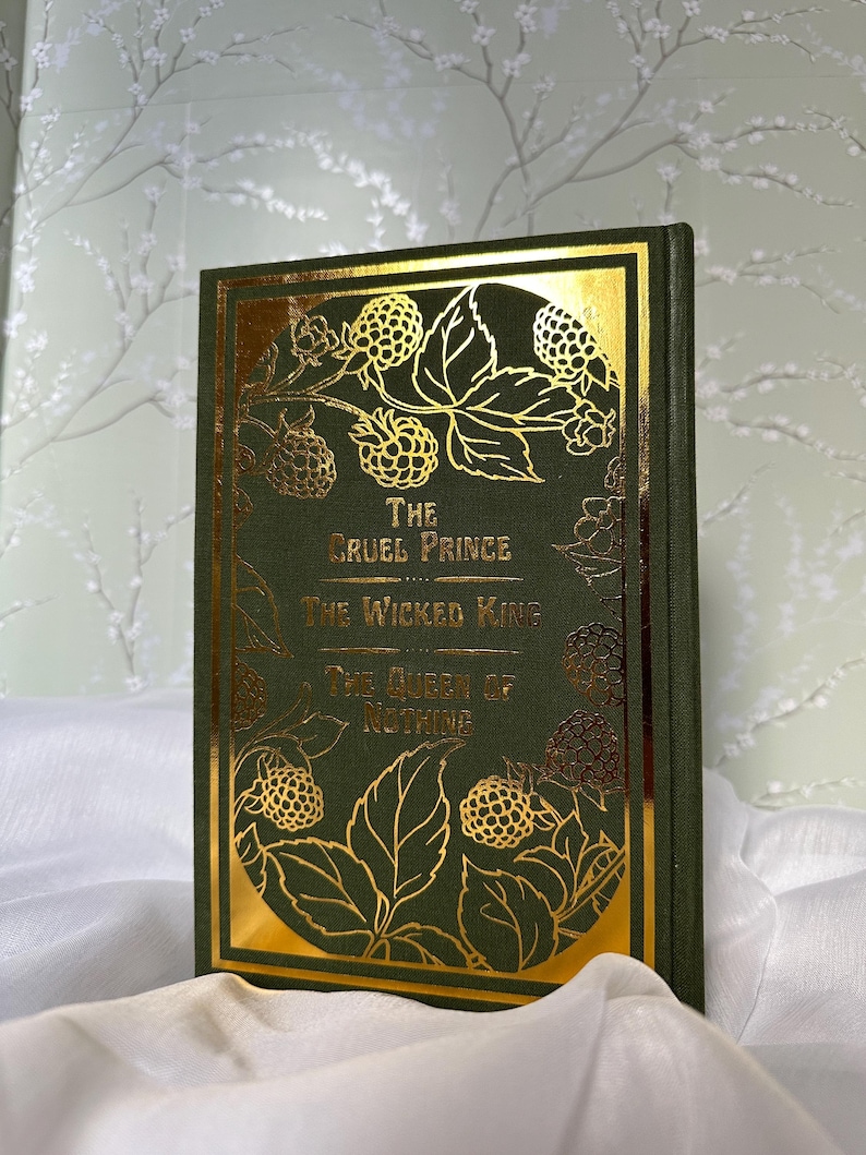 May include: A hardcover book with a dark green cover and gold foil accents. The title "The Cruel Prince," "The Wicked King," and "The Queen of Nothing" are displayed in gold lettering. The cover features gold illustrations of berries and leaves.