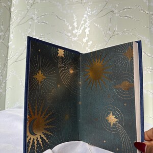 May include: Open book with a navy blue cover and gold celestial illustrations. The interior pages feature a dark teal background with gold suns, stars, and planets. The book is open, displaying the detailed artwork.