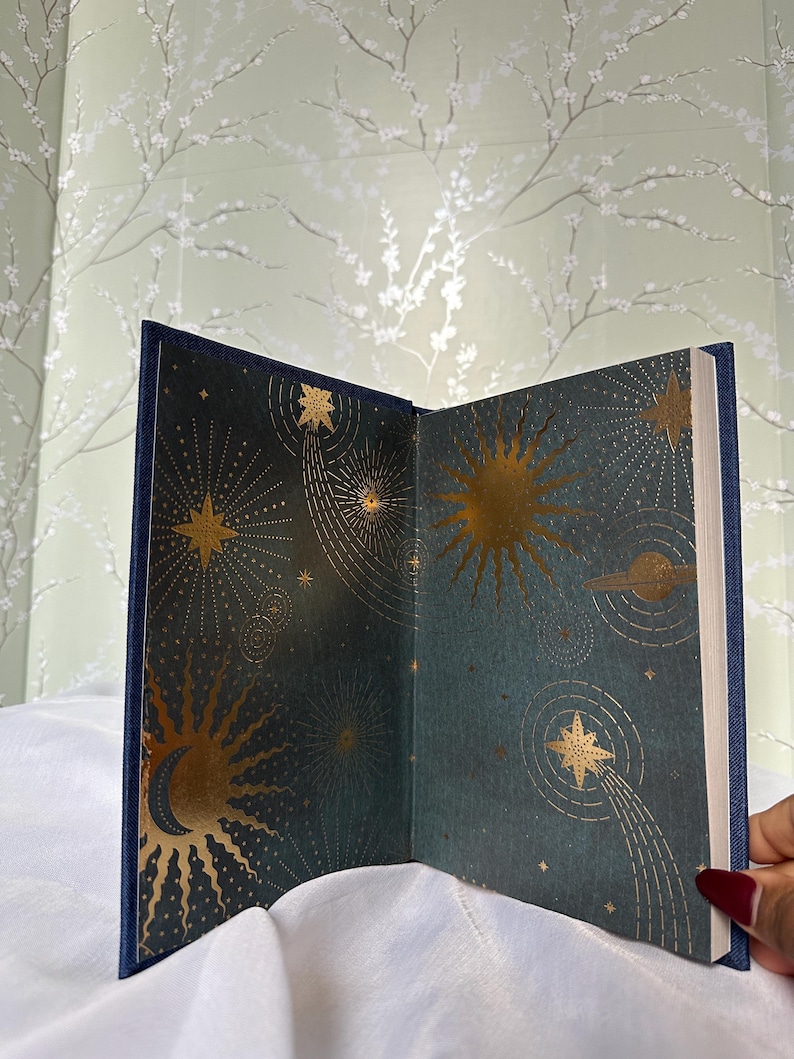 May include: Open book with a navy blue cover and gold celestial illustrations. The interior pages feature a dark teal background with gold suns, stars, and planets. The book is open, displaying the detailed artwork.
