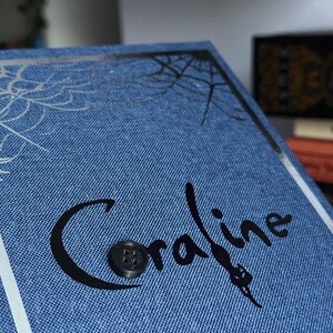 May include: A blue book with the title "Coraline" in black script, featuring a button and a spiderweb design. The book has a denim-like texture and a white border.