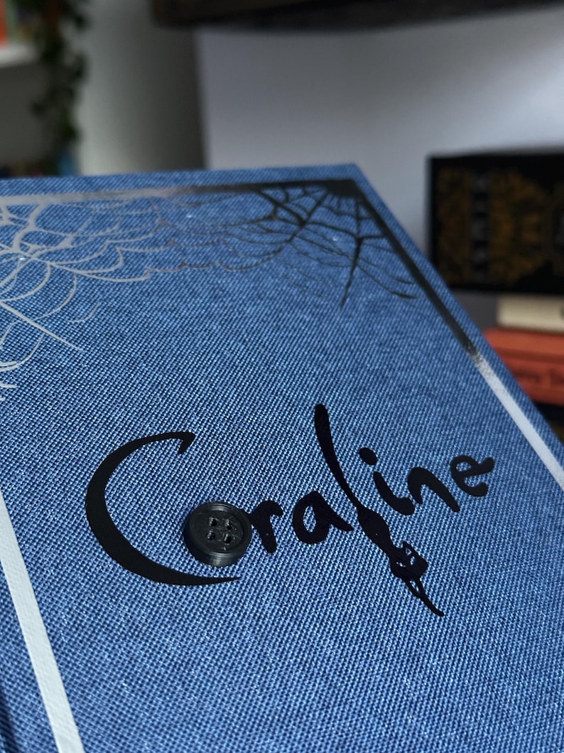 May include: A blue book with the title "Coraline" in black script, featuring a button and a spiderweb design. The book has a denim-like texture and a white border.