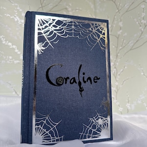 May include: A navy blue book titled "Coraline" with silver spiderweb and spider accents. The book has a textured cover and a silver border. The title is in a stylized font with a button detail.