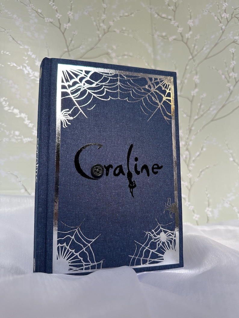 May include: A navy blue book titled "Coraline" with silver spiderweb and spider accents. The book has a textured cover and a silver border. The title is in a stylized font with a button detail.