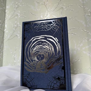 May include: A dark blue book with a silver illustration of a figure in a tunnel. The cover features a black frame with spiderwebs and spiders. The book is on a white cloth.