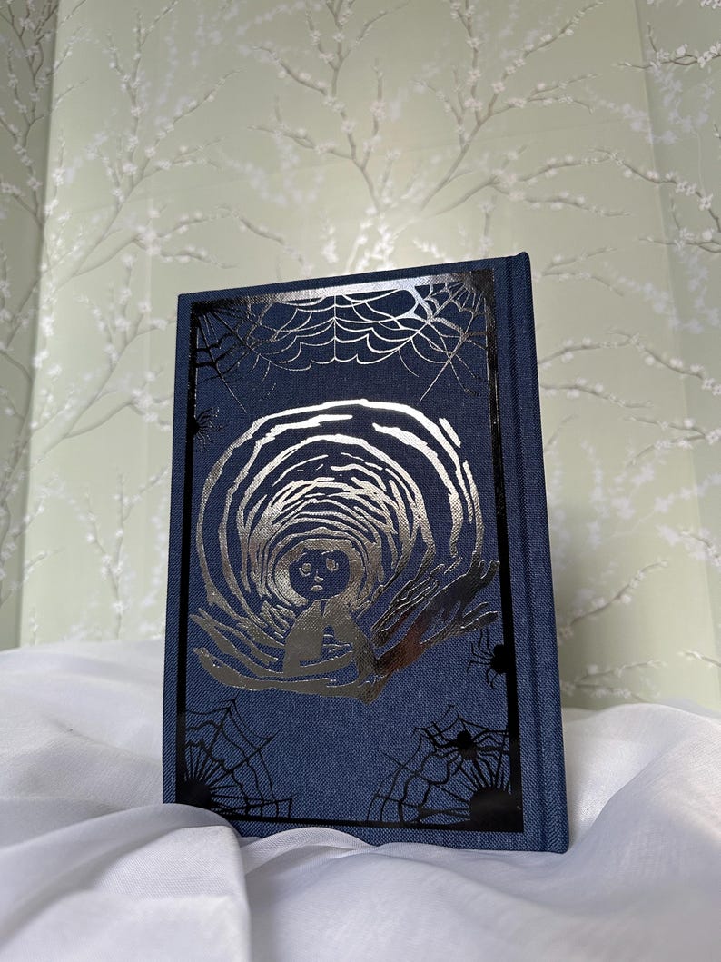 May include: A dark blue book with a silver illustration of a figure in a tunnel. The cover features a black frame with spiderwebs and spiders. The book is on a white cloth.