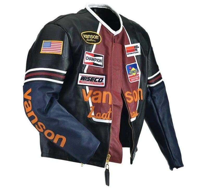Peut inclure: Veste de moto en cuir noir avec un panneau central bordeaux et des manches bleues. La veste comporte plusieurs &eacute;cussons, dont un drapeau am&eacute;ricain, et les mots "Vanson Leathers" en orange.