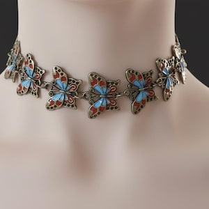 May include: A bronze-toned choker necklace featuring butterfly pendants. Each butterfly has red, blue, and white enamel details. The necklace is displayed on a mannequin.