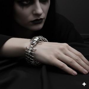 Könnte beinhalten: Ein silberfarbenes Armband in Form eines Tausendfüßlers wird am Handgelenk getragen. Das Armband hat segmentierte Körperabschnitte und mehrere Beine. Der Hintergrund ist dunkel, mit einer Person in schwarzer Kleidung und dunklem Make-up.