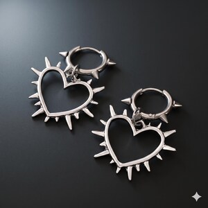 May include: A pair of silver-colored hoop earrings with a heart-shaped pendant. The heart pendant is outlined with spikes. The hoop earrings have three spikes. The earrings are displayed on a dark gray surface.