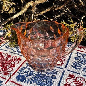 May include: A vintage pink glass creamer with a geometric diamond pattern. The creamer has a spout and a handle. It sits on a patterned tablecloth with red and blue floral designs. The background includes branches and sunlight.