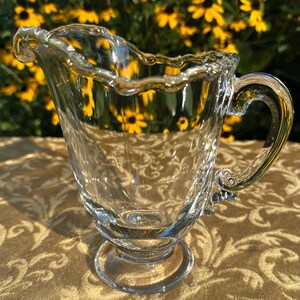 Puede incluir: Una jarra de cristal transparente para crema con borde festoneado, asa curva y base circular con pedestal. La jarra tiene una superficie lisa y transparente con un diseño moldeado que crea sutiles estrías verticales en el cuerpo.
