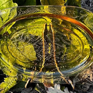 May include: An oval-shaped, antique glass serving platter with a decorative floral pattern. The platter is a pale yellow-green color with a brown rim and handles. The edges have a detailed, embossed design.
