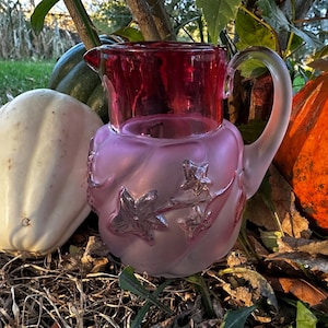 May include: A vintage glass pitcher with a ruby red top and a frosted pink base. The base is decorated with star and leaf designs. The pitcher has a curved handle and is set among pumpkins and gourds, suggesting a fall theme.