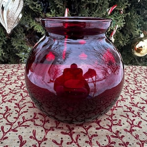 May include: A round, ruby red glass vase with a wide opening. The vase reflects the surrounding environment, including a Christmas tree with ornaments. The vase sits on a patterned surface with a red and beige design.