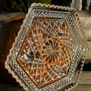 May include: A clear, hexagonal glass dish with a detailed diamond pattern. The dish is resting on a pumpkin, creating a warm, autumnal aesthetic. The intricate design of the glass catches the light, adding a touch of elegance.