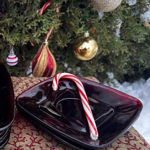 Puede incluir: Una pila de platos cuadrados de vidrio rojo oscuro con un bastón de caramelo encima. Los platos están sobre una tela estampada con un árbol de Navidad y adornos en el fondo.