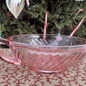 May include: A pink glass bowl with a handle, featuring a swirled design. The bowl is set on a patterned surface with Christmas decorations in the background, including candy canes and ornaments.