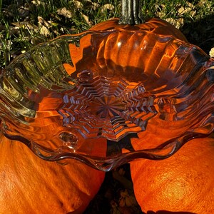 Könnte beinhalten: Eine klare Glasschüssel mit Sternmuster in der Mitte liegt auf drei leuchtend orangefarbenen Kürbissen. Die Schüssel hat einen wellenförmigen, gewellten Rand. Die Kürbisse sind in verschiedenen Größen und Formen mit grünen Stielen.