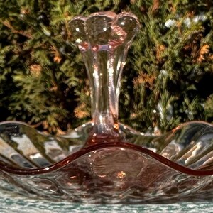 Puede incluir: Un plato de servir vintage de cristal rosa con un asa central. El plato tiene un borde festoneado y una superficie texturizada. El asa tiene un diseño decorativo y acanalado. El plato se utiliza probablemente para servir aperitivos o postres.