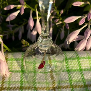 May include: A clear glass bell with a faceted handle and a smooth, rounded body. A small, deep red, diamond-shaped crystal charm hangs from a thin gold-toned metal chain attached to the interior top of the bell, serving as the clapper.