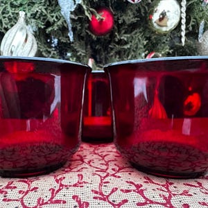 Può includere: Due tazze di vetro rosso rubino con un motivo strutturato sulla base, appoggiate su una tovaglia fantasia. Un contenitore di vetro rosso più piccolo è tra le tazze. Lo sfondo presenta un albero di Natale con ornamenti.