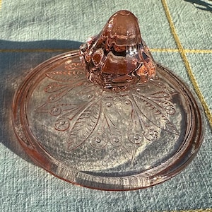 Könnte beinhalten: Vintage rosa Glasdeckel mit dekorativem Knauf. Der Deckel hat ein geätztes Pfauenfedermuster. Der Knauf hat eine abgerundete Form. Das Glas hat einen leichten rosa Farbton und ist transparent.