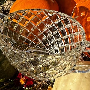 Könnte beinhalten: Eine klare, diamantgeschliffene Glasservierschale mit Griffen. Die ovale Schale steht zwischen Kürbissen und Zierkürbissen und reflektiert die orangefarbenen und grünen Farbtöne der Erntezeit. Das Glas funkelt im Sonnenlicht.