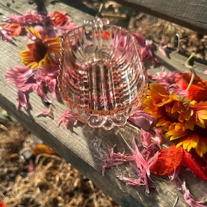 May include: A pale pink pressed glass dish featuring a vertical ribbed pattern along the sides and a scalloped rim. The oval-shaped vessel includes small, decorative scroll-like handles on each end, showcasing a translucent and reflective finish.