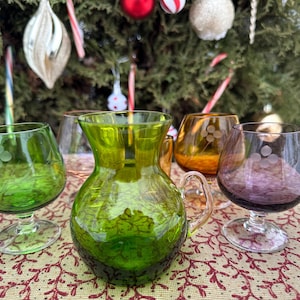 May include: A selection of vintage green, amber, and purple glass drinkware. Includes a jug with a handle and several stemmed glasses. The glasses have etched dot patterns. The background features a Christmas tree with decorations and candy canes.