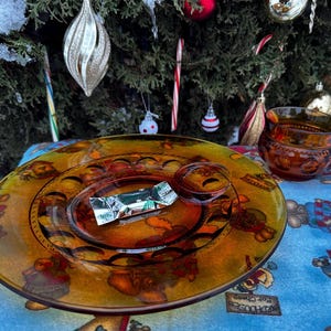May include: A clear amber glass plate with a candy wrapper on it, and a matching cup. The plate has a decorative pattern around the edge. The items are on a blue tablecloth with a Christmas tree in the background.