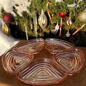 Puede incluir: Cinco fuentes de vidrio rosa con forma triangular y diseño acanalado. Los platos están dispuestos sobre una superficie marrón, con un fondo borroso de adornos navideños y vegetación.