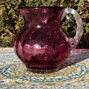 May include: A hand-blown, ruby-colored glass pitcher with a clear, twisted handle. The pitcher has a rounded body and a flared spout. The glass has a textured, crackled appearance, and the pitcher sits on a patterned surface.